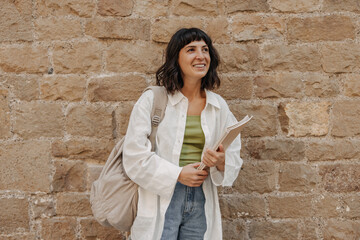 Wall Mural - Pretty smiling woman looking away at the street. European brunette student holding noteeboks, with backpack on the wall background. Positive emotion concept