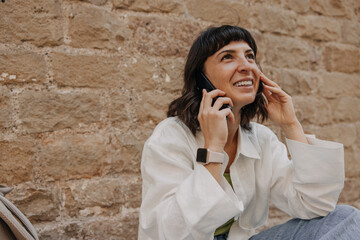 Wall Mural - Picture of charming caucasian woman talking on cellphone at the street. Dark-haired lady in white t-shirt on the wall background. Telephone technology concept