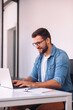 © kerkezz - A smiling man with glasses is using a laptop in the office while sitting at his desk.