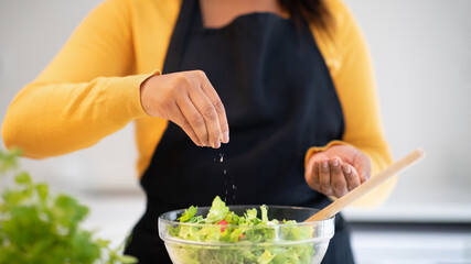 Wall Mural - Millennial african american woman in apron salting salad, cooking dinner at table with organic fresh vegetables