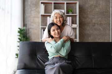 Loving adult daughter hugging older mother, standing behind couch at home, family enjoying tender moment together, young woman and mature mum or grandmother looking at each other, two generations