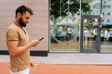 Side view of confident young man in casual clothes walking on city street, using mobile phone and drinking takeaway coffee on summer day. Bearded hipster male typing smartphone outdoors.