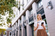 © dikushin - Low-angle view of positive blonde young woman in casual clothes drinking takeaway coffee to go and using smartphone walking on urban street, smiling looking away. Coffee break concept.