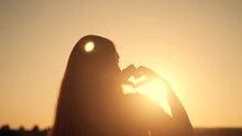 Happy Girl In Park At Sunset. Finger-shaped Heart Shape. Hands Of Girl Shape Of Heart. Summer Dream. Happiness Of Freedom In A Field At Sunset.Sunlight Between Fingers.Silhouette Of Happy Girl In Park
