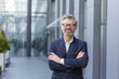 © Liubomir - Portrait of mature gray-haired businessman, senior boss smiling and looking at camera with arms crossed, gray-haired man outside office building, successful investor and owner in business suit.