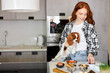 © Roman - Redhead Lady In Casual Wear Is Preparing Meal Food For Pet Dog, Feeding. Fresh Food On Table In Kitchen At Home, Woman Holding Dog In Hands, Caring And Loving. People, Animals, Lifestyle Concept