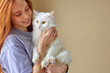© Roman - Pets Care. redhead caucasian woman in casual wear holding cat in hands hugging, at home. Friendship.Love. adorable owner with natural red hair with cute purebred white cat. portrait