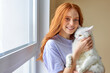 © Roman - Portrait Of Young Redhead Woman With White Cat In Hands Posing At Camera, At Home, Smiling Happily. At Weekends, Holidays. Sweet Adorable Cat With White Fur is Relaxed In Hands Of Owner