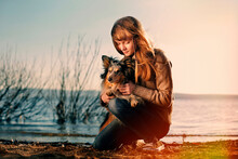 Teenage Girl Holding Dog While Kneeling On Beach During Sunset