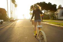 Young Woman Wearing A Headphone, Yellow Socks And A Hoodie Rides A Cruiser Bike To The Beach. (backlite)