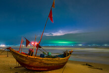 A Fishing Boat Flying Red Flags Sitting High On A Beach At Night With A Lightning Storm Behind At Mui Ne In Vietnam