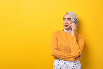 Beautiful pensive Asian girl wearing hijab talking on cell phone, serious face thinking question isolated on yellow background