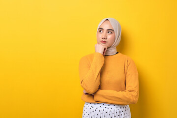 Wall Mural - Beautiful pensive Asian girl wearing hijab looking aside at empty space, touching chin, thinking about offer isolated on yellow background