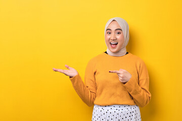 Wall Mural - Beautiful excited Asian girl wearing hijab pointing finger at open palm, advertising something isolated on yellow background