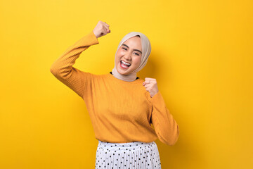 Wall Mural - Beautiful excited Asian girl wearing hijab raising fist, celebrating success isolated on yellow background