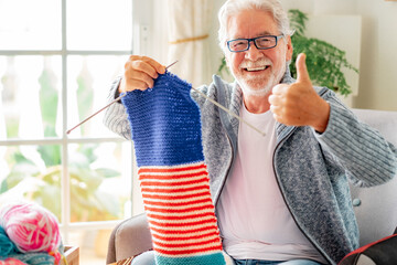 Canvas Print - Portrait of happy senior man proud of his knitted wool sitting on the armchair at home. Elderly caucasian man knits wool looking at camera with thumb up.