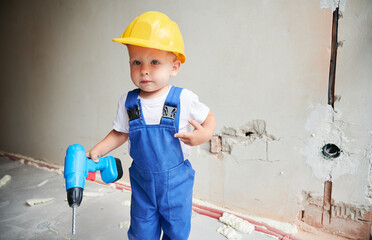 Child with toy construction tool for repair works standing against the wall in apartment under renovation. Kid in safety helmet and work overalls holding electric drill and looking at camera.