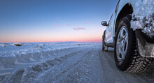Perspective Wheel Of 4x4 Car With Ice On A Completely Snowy Road With Tire Tracks That Goes To The Horizon With A Golden And Magical Icelandic Sunrise. The Light Of Dawn Is Reflected In The Car.
