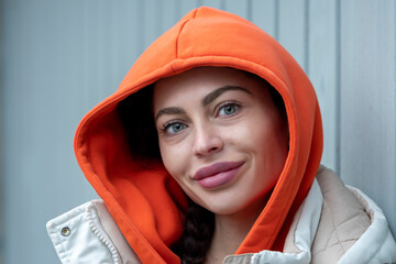 A beautiful 25-30-year-old woman in a jacket and an orange hood is looking at the camera, leaning against metal shutters.