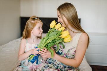 Wall Mural - Cute little girl greeting mother and gives her a bouquet of yellow flowers tulips at home. Mother's day concept. Mom and daughter smiling and kissing. Happy family holiday and togetherness. Side view.