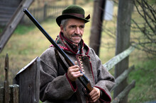  A Hunter With A Gun In His Hands In Hunting Clothes Stands Near The Forest Hut.