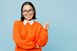 © ViDi Studio - Young smiling happy woman of Asian ethnicity wearing orange sweater glasses point index finger aside indicate on workspace area copy space mock up isolated on plain pastel light blue cyan background.