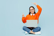 © ViDi Studio - Full body fun young IT woman of Asian ethnicity wear orange sweater glasses hold use work on laptop pc computer sitting point finger on area isolated on plain pastel light blue cyan background studio.