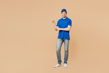 Wall Mural - Full body smiling delivery guy employee man wear blue cap t-shirt uniform workwear work as dealer courier point index finger aside on area isolated on plain light beige background. Service concept.