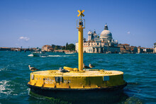 Floating Yellow Buoy In The Lagoon Of Venice And The Turquoise Sea Against The Blue Sky And Ancient Venetian Houses, Seagulls Rest On A Mooring Barrel In The Grand Canal Of Venice, Tourist Boats
