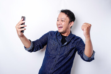 A young Asian man with a happy successful expression wearing blue shirt and holding his phone, isolated by white background