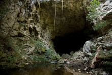 A Traveller Walks On The River Rocks Beneath A Giant Cave Opening.