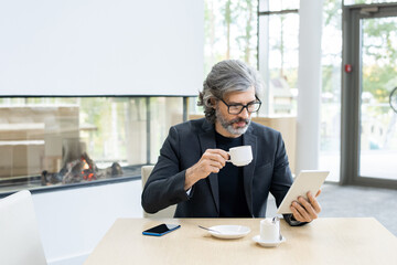 Wall Mural - Bearded mature economist or banker in suit and eyeglasses analyzing online financial data in tablet by cup of coffee in modern restaurant