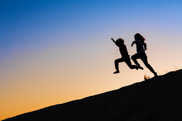 Wall Mural - Black silhouette at sunset sky background. Happy family - kids running and jumping high in the air from sea beach sand dune. Active people, outdoor activity on tropical summer vacations with children