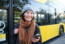 Portrait Of Girl Standing Near Bus On A Stop, Waiting For Her Public Transport, Schecks Schedule On Smartphone Application, Holds Mobile Phone, Wears Warm Clothes