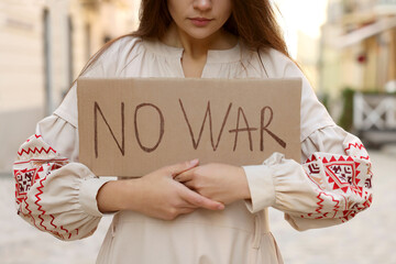 Wall Mural - Sad woman in embroidered dress holding poster No War on city street, closeup