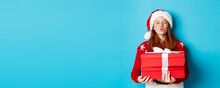 Happy Holidays And Christmas Concept. Cute Redhead Girl Holding Presents And Pucker Lips For Kiss, Wearing Santa Hat And Funny Sweater, Blue Background