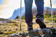 Legs Of A Hiker In Trekking Boots Walking In The Mountains With Nordic Walking Poles Closeup Shot. Feet Of Walking Tourist Wearing Trekking Shoes With Hiking Sticks On Rocky Road Captured From Behind