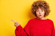Portrait of emotional curly haired woman, indicates left to the copy space, has positive expression, demonstrates something amazing, dressed in bright clothes over yellow background