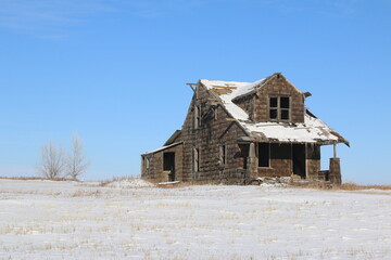 Outdoor winter landscape of an old abandoned wooden two story house on the empty prairie under a clear blue sky.