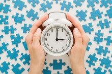 White Alarm Clock In Young Adult Woman Palms. Different Many Blue Puzzle Pieces On Table Background. Closeup. Spending Time Concept. Point Of View Shot. Top Down View.