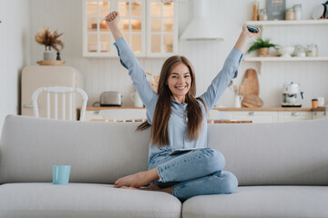 Attractive blonde caucasian woman in casual sits on sofa at home eyes closed broad smiles rises hands up in winner expression against blurry kitchen. Beautiful Italian girl got great news. Cosy home.