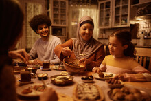 Happy Muslim Mature Woman Serving Food To Her Family During Meal At Dining Table.