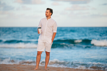 Wall Mural - Young man in white with coffee on the beach walking and enjoy evening time