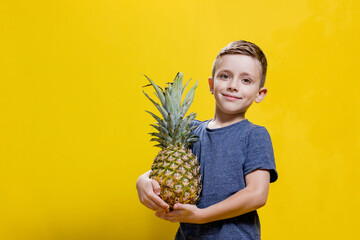 Wall Mural - Child boy holding pineapple while posing on yellow background with copy space. Concept of summer and healthy vegan food