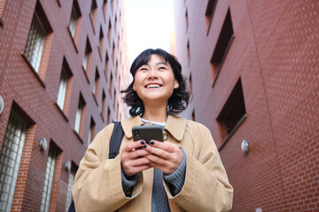 Wall Mural - Lower angle view of brunette korean girl, listening music in headphones, walking along street and looking at smartphone, reading message on mobile phone