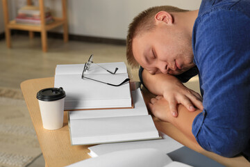 Wall Mural - Tired man sleeping near books at wooden table indoors