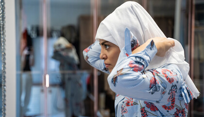 Wall Mural - Portrait of middle-aged muslim woman getting dressed hijab head scarf in fitting room  looking at mirror. Side view face of cheerful woman covered with headscarf. Casual islamic girl. Copy space