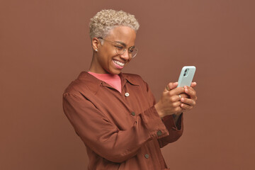 Young positive beautiful African American woman wearing glasses holds phone and be touched seeing interesting post on social networks or reading story with happy ending stands in brown studio