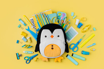 Wall Mural - Plush penguin backpack with stationery and supplies for drawing and craft on yellow background. Various colorful material for creativity and art activity. Primary School or kindergarten.