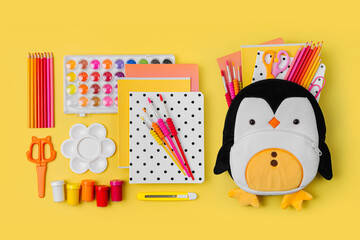 Wall Mural - Plush penguin backpack with stationery and supplies for drawing and craft on yellow background. Various colorful material for creativity and art activity. Primary School or kindergarten.